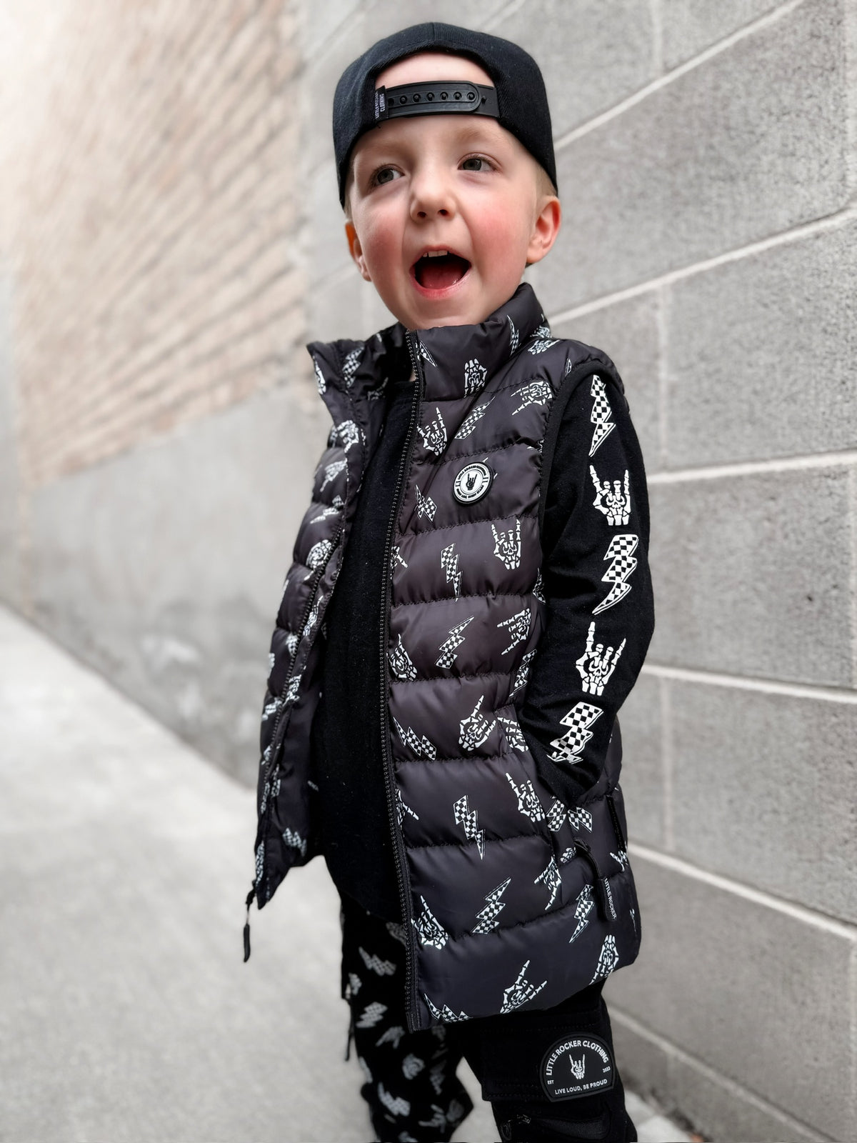 Kid wearing Kids black puffer vest with Bolted Bones design featuring skeleton hand rock on graphic and lightning bolts, rocker style outerwear for kids