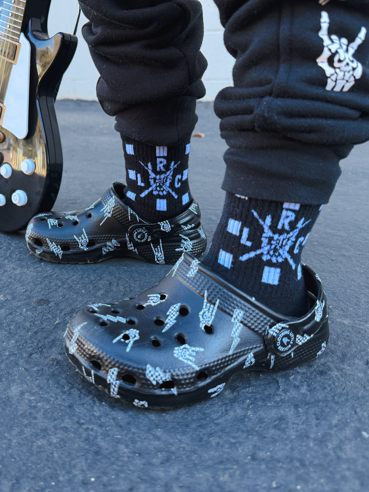 Black bolted bone kinsley clogs and black crew socks with a little rocker clothing design, worn by a person standing outdoors on asphalt next to an electric guitar.