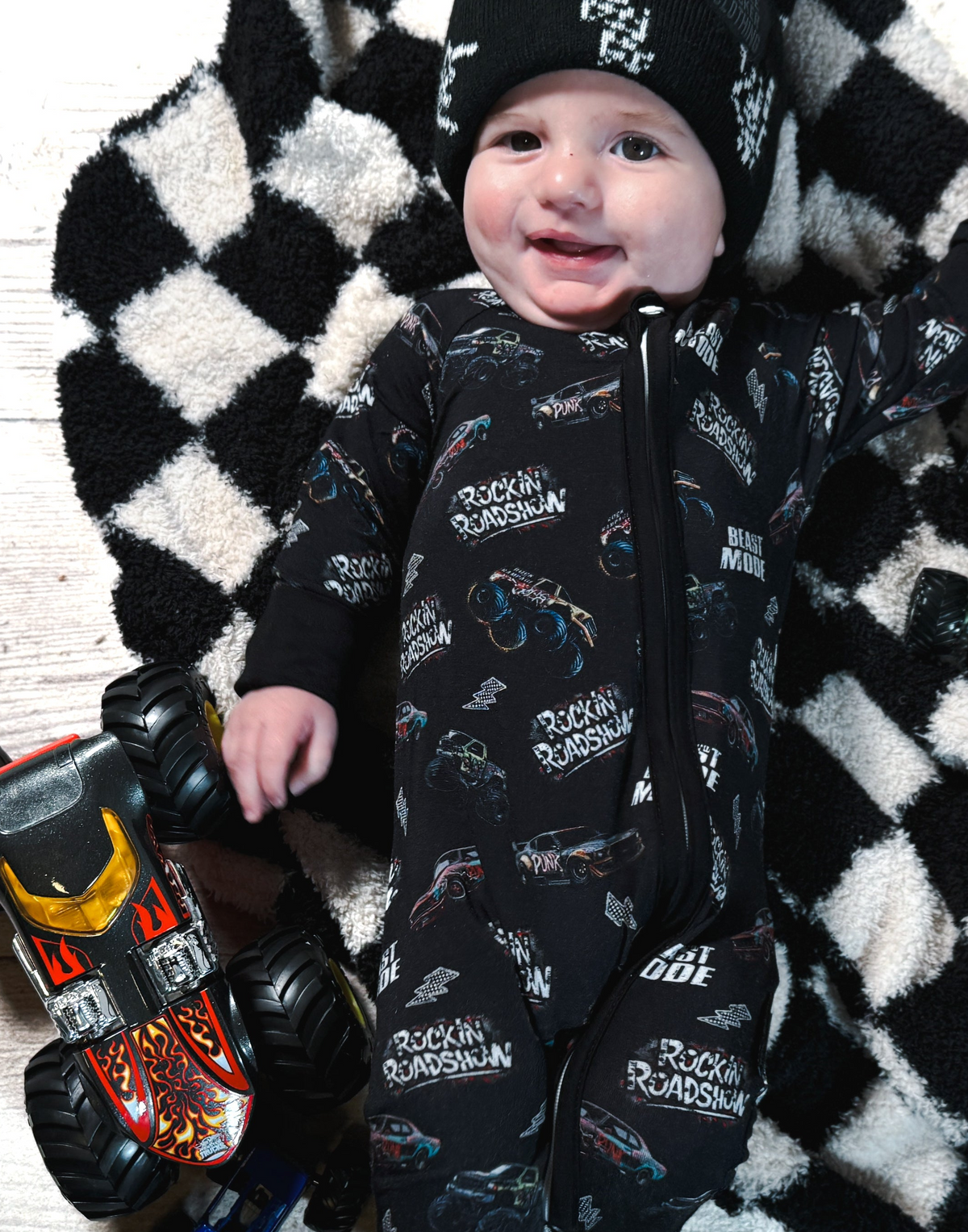 infant wearing black bamboo pajamas with a monster truck pattern while laying on a checkered rug