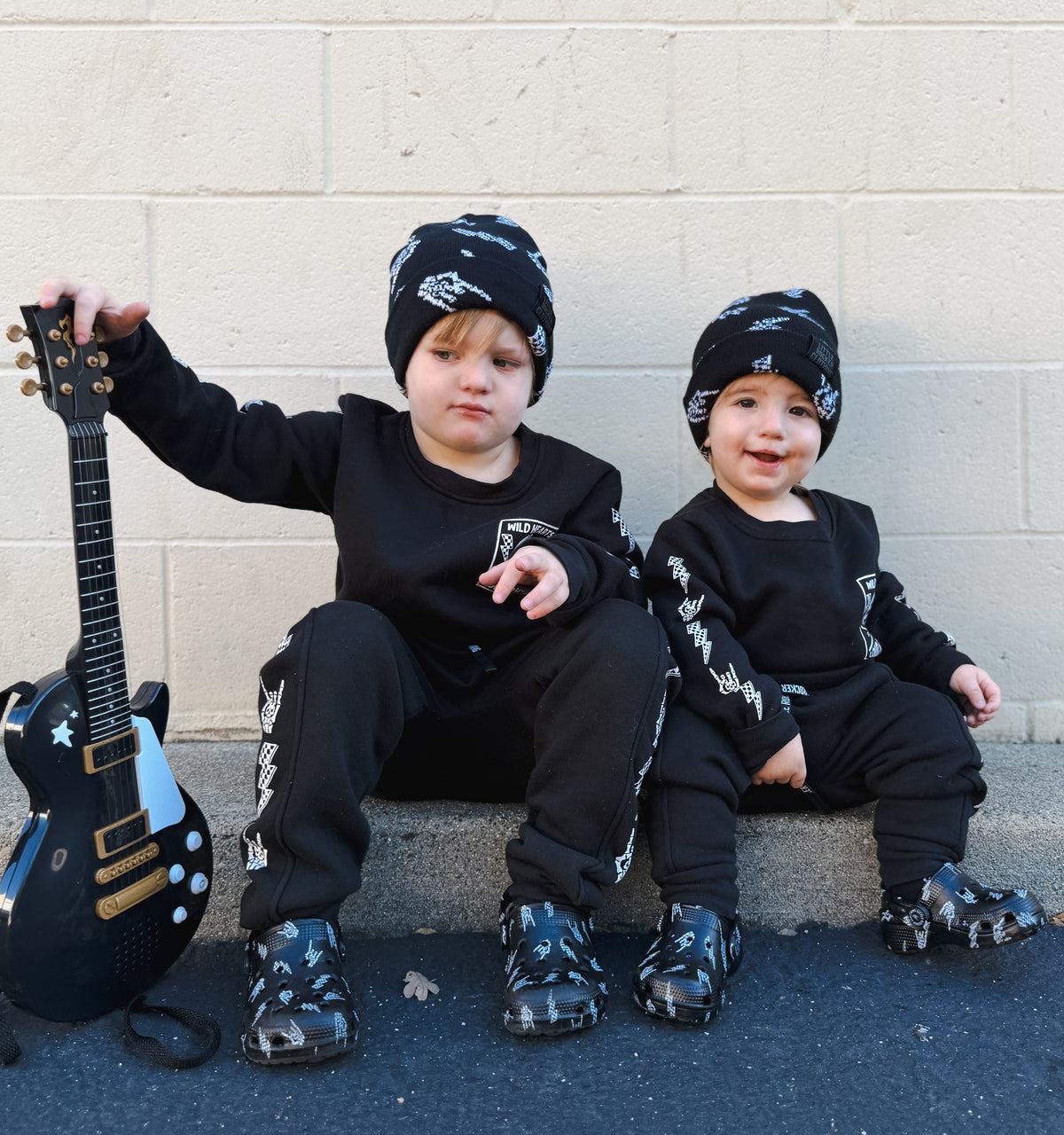 Two toddlers sitting on a concrete ledge against a light beige brick wall, wearing matching black long-sleeve sweatshirts, black pants with white graphic prints, and black beanie hats from little rocker clothing. One toddler holds a black electric guitar toy.