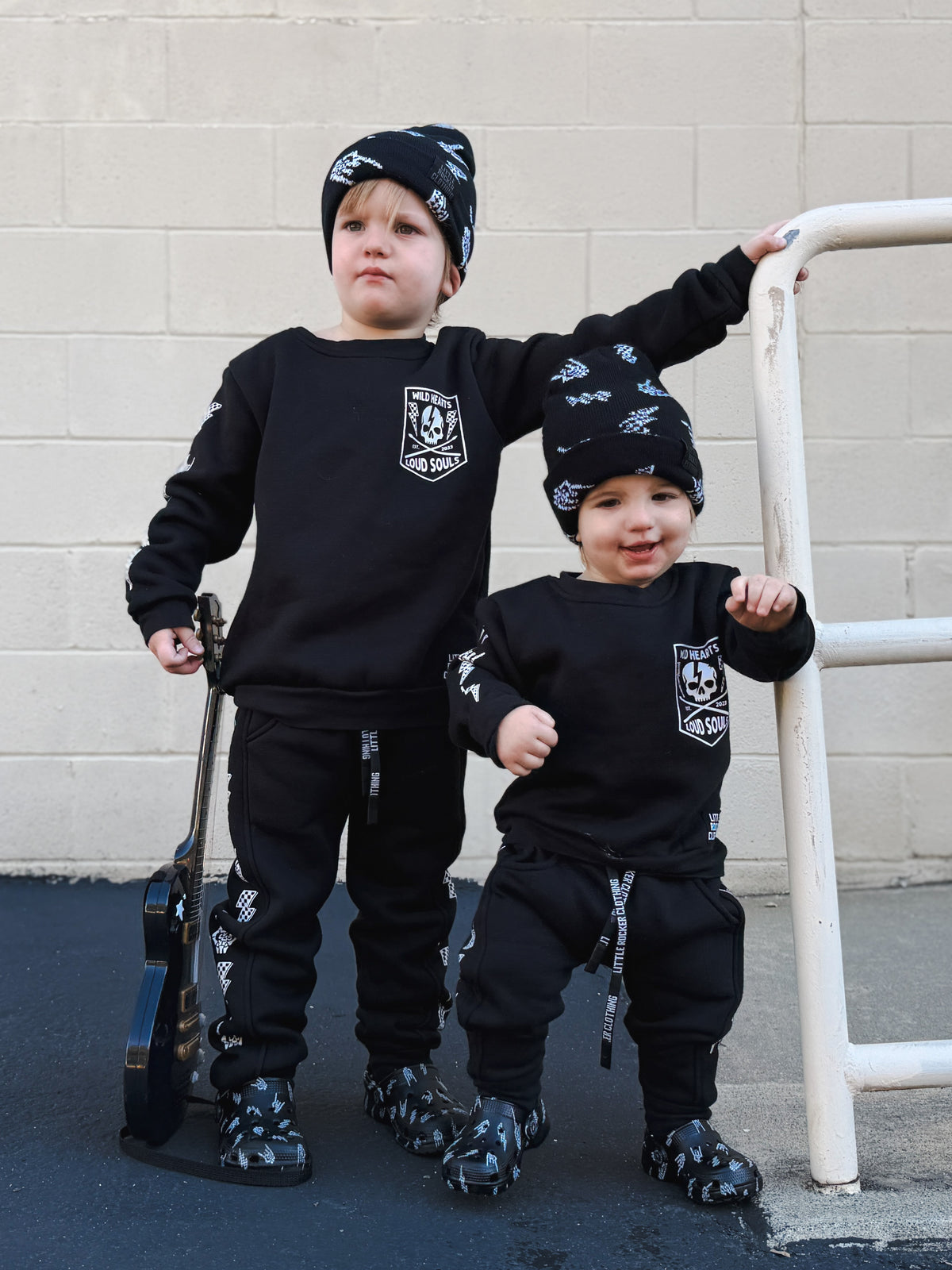 Two toddlers wearing matching black outfits with white graphic prints, standing outdoors against a light gray brick wall. One child holds a toy guitar, and both wear patterned black-and-white beanies and black pants from little rocker clothing.