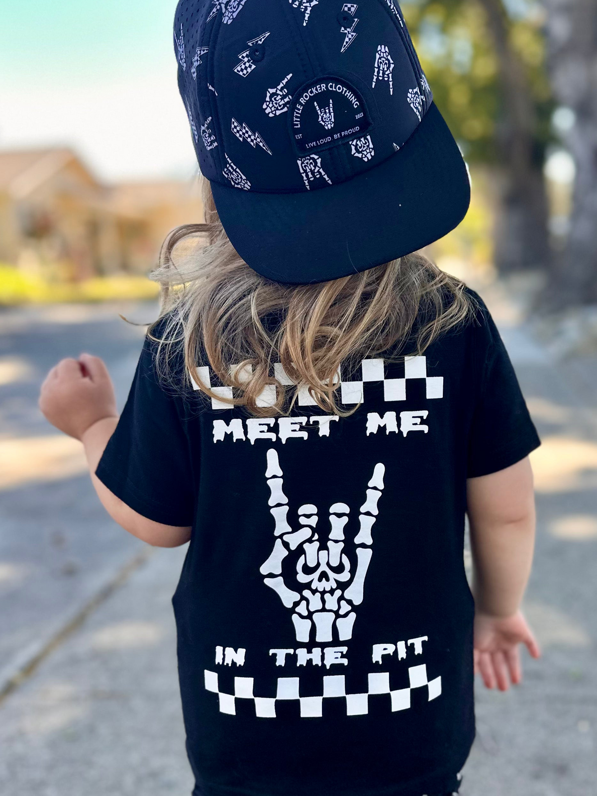 black kids graphic t-shirt with skeleton hand rock on design and “meet me in the pit” text, checkerboard detail, little rocker clothing rock style outfit for toddlers and kids outdoors rear view