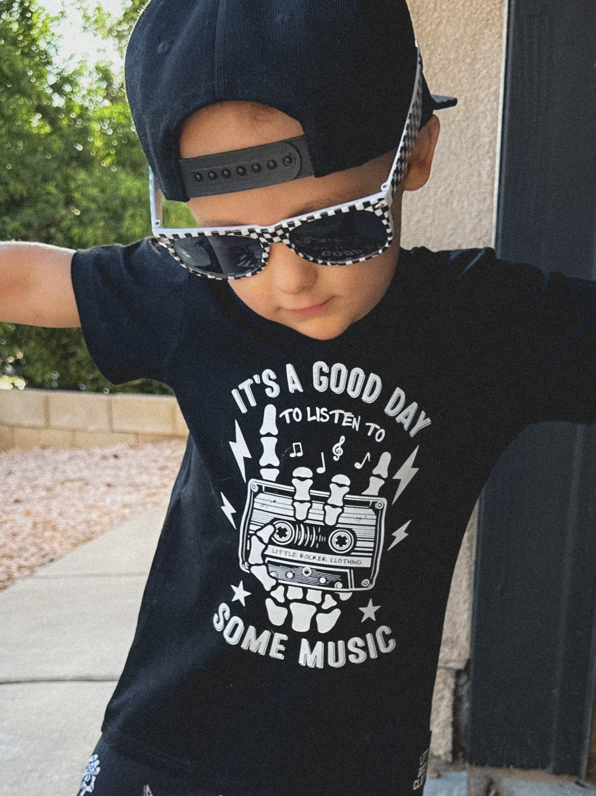 black kids graphic t-shirt with skeleton hands holding cassette tape and “it’s a good day to listen to some music” text, little rocker clothing rock style outfit with checkered sunglasses and snapback front view