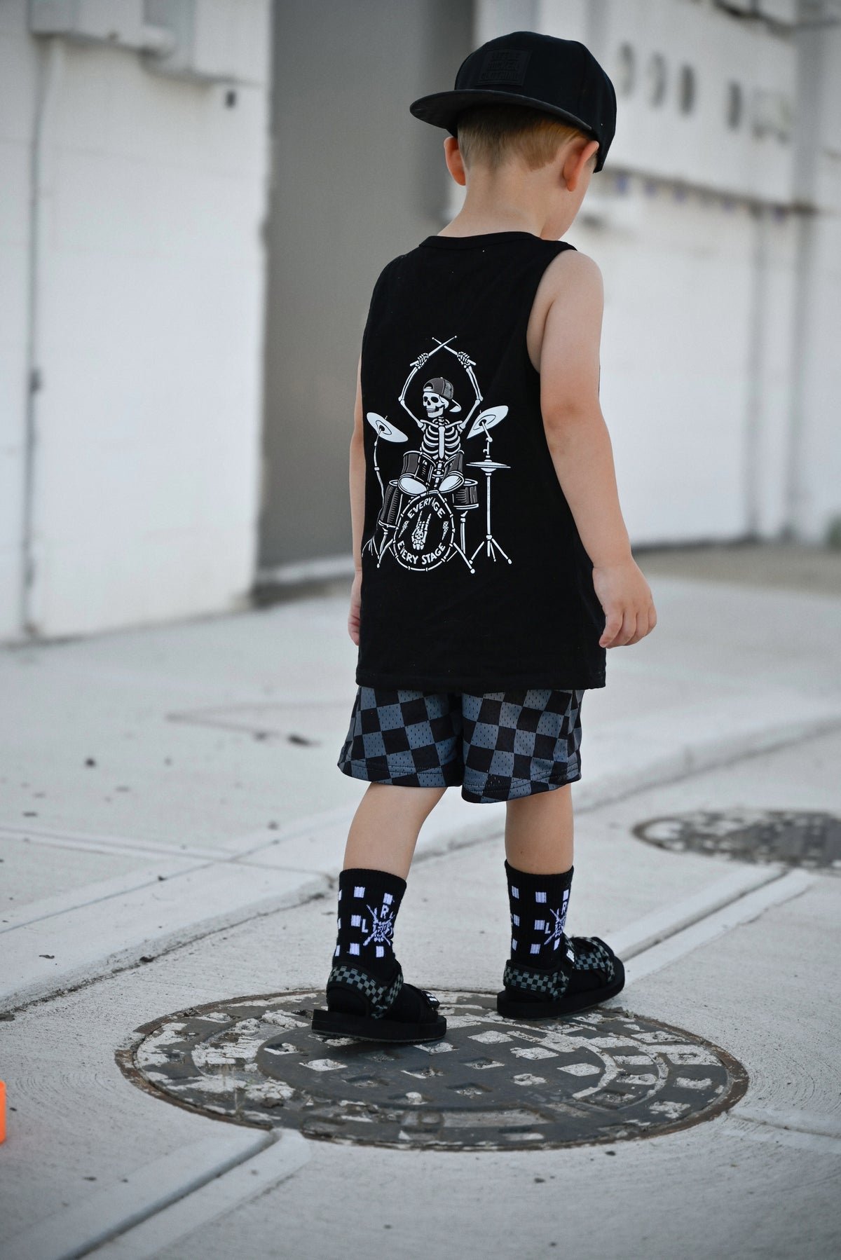 child wearing backwards snapback hat, drumming skeleton tank top and checkered shorts from little rocker clothing
