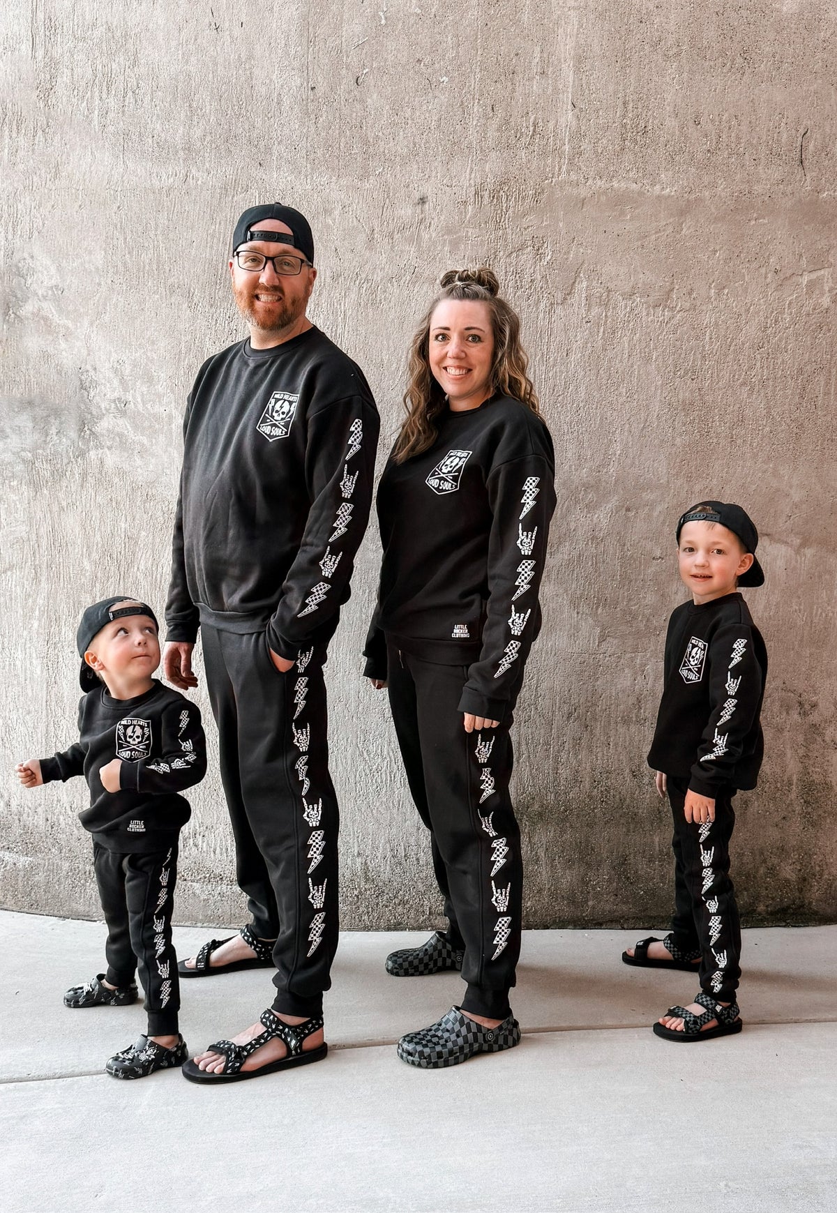 family wearing matching black aftershock crew neck sweatshirts and joggers set lightning bolt and rock hand print little rocker clothing adult and kids outfits