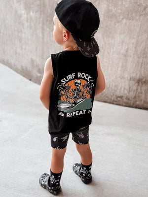 Toddler wearing black Little Rocker Clothing tank top with skeleton surfing graphic and “Surf Rock Repeat” design, paired with black snapback hat
