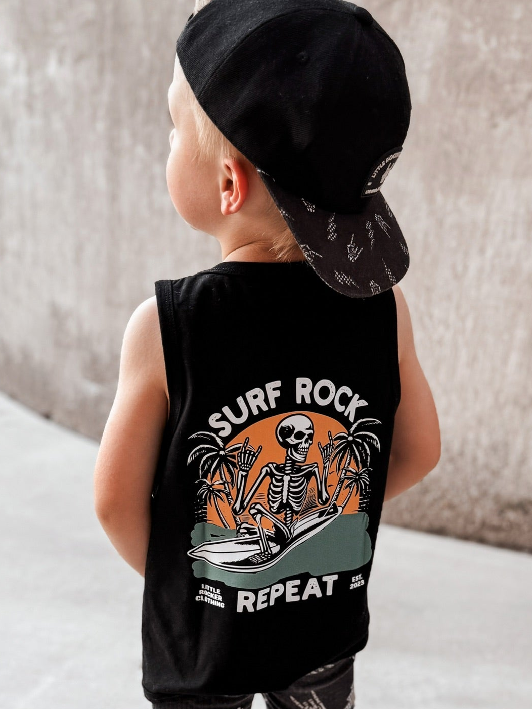 Toddler wearing black Little Rocker Clothing tank top with skeleton surfing graphic and “Surf Rock Repeat” design, paired with black snapback hat