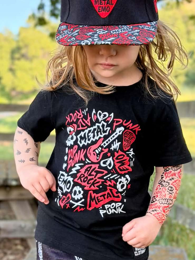 A toddler wearing a black graphic t-shirt with punk rock and metal inspired lettering, matching black shorts with white skull prints, a black cap, and slip-on shoes, standing outdoors in front of a white wooden wall.
