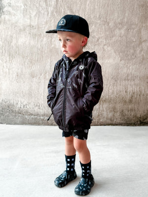 Young boy wearing the Kids bolted bones black windbreaker jacket with all over graphic print, live loud be proud on back side of the lightweight rocker style outerwear for kids with zip front and hood