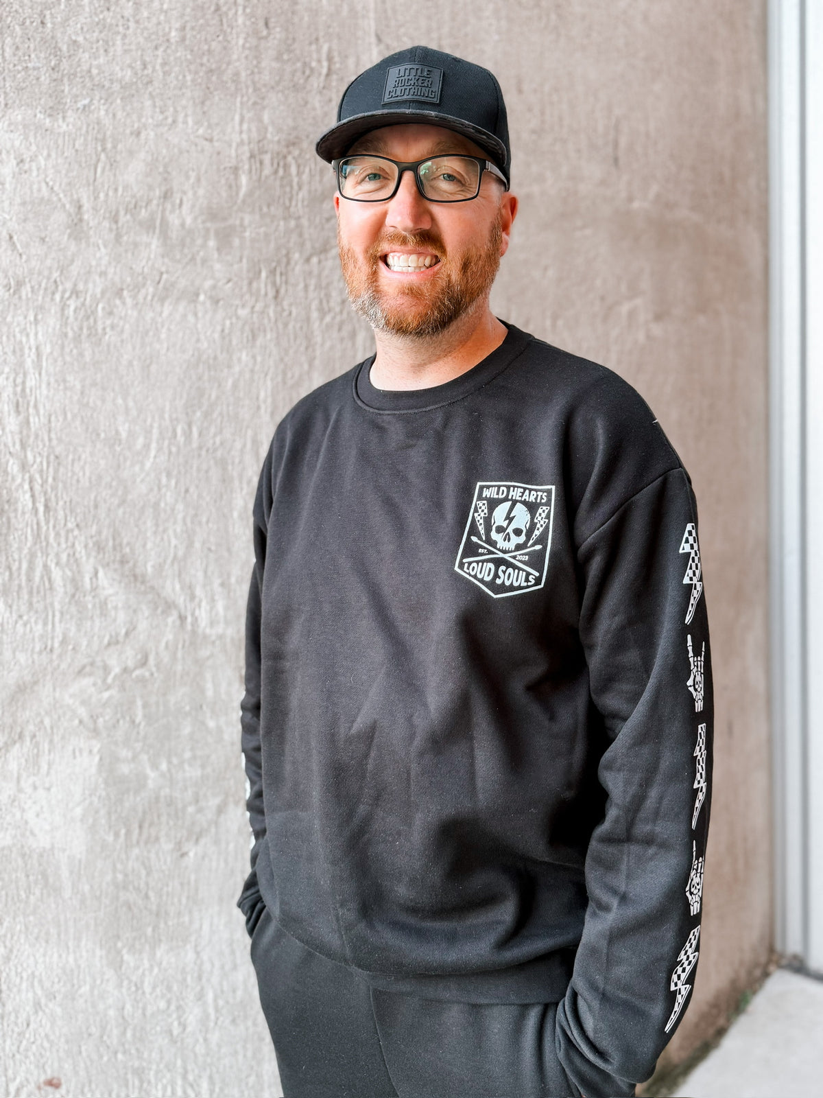 adult man wearing matching black aftershock crew neck sweatshirts and joggers set lightning bolt and rock hand print little rocker clothing adult and kids outfits