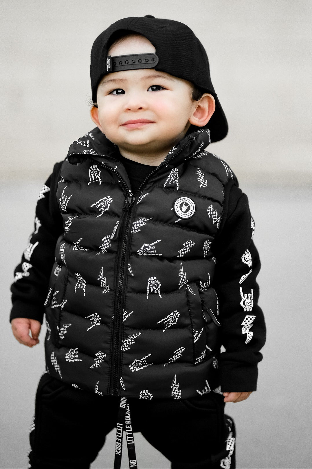 Infant wearing Kids black puffer vest with Bolted Bones design featuring skeleton hand rock on graphic and lightning bolts, rocker style outerwear for kids