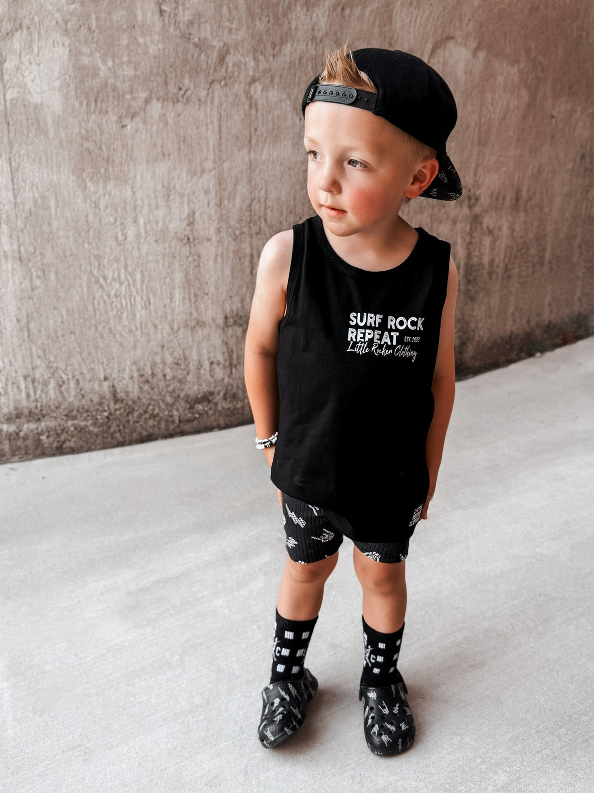 Toddler boy wearing black Little Rocker Clothing tank top with skeleton surfing graphic and “Surf Rock Repeat” design, paired with black snapback hat