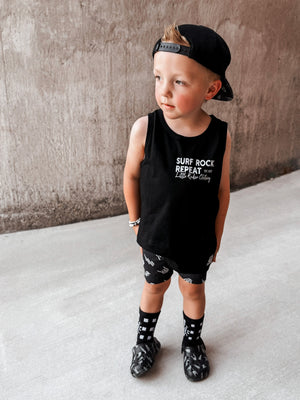 Toddler boy wearing black Little Rocker Clothing tank top with skeleton surfing graphic and “Surf Rock Repeat” design, paired with black snapback hat