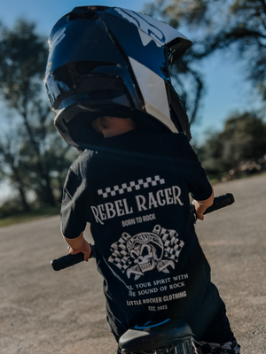 black kids “rebel racer” graphic t-shirt with skull and checkered flag design and “born to rock” text, little rocker clothing outfit worn by child riding bike with helmet, rear view outdoors