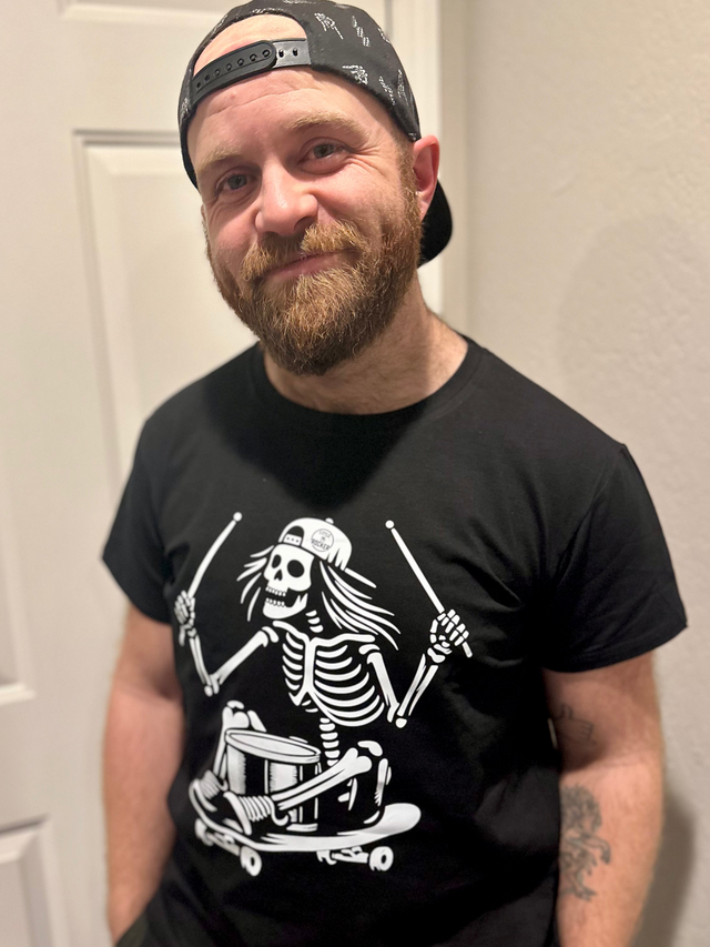 adult man wearing a black graphic tshirt featuring a skeleton sitting on a skateboard with drumsticks 