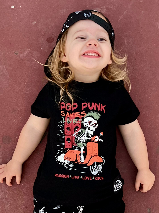 black kids graphic t-shirt with “pop punk saves lives” skeleton riding scooter design in red and white, little rocker clothing punk rock toddler outfit front view smiling child outdoors