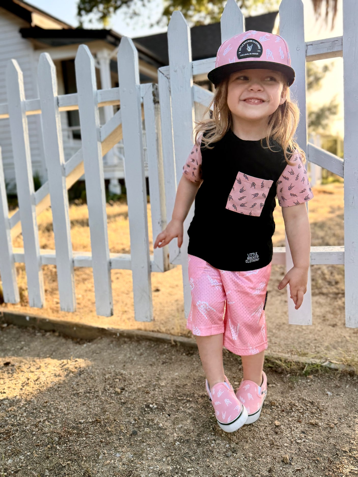 A young girl smiles while wearing a Little Rocker Clothing coordinating outfit: a black raglan tee with a pink patterned pocket and sleeves, matching pink patterned shorts, a pink baseball cap, and pink slip-on sneakers. She stands on a gravel path in front of a white wooden fence.