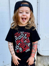 A toddler wearing a black graphic t-shirt with punk rock and metal inspired lettering, matching black shorts with white skull prints, a black cap, and slip-on shoes, standing outdoors in front of a white wooden wall.
